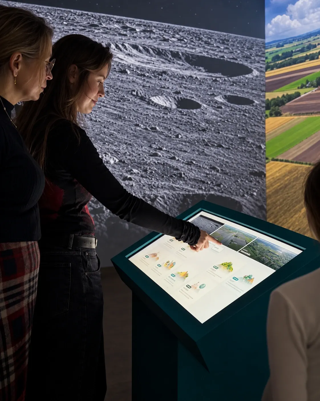 Enhanced video still of visitors using Vignette's touchscreen controller beside immersive wall projections of cratered terrain and a green landscape.